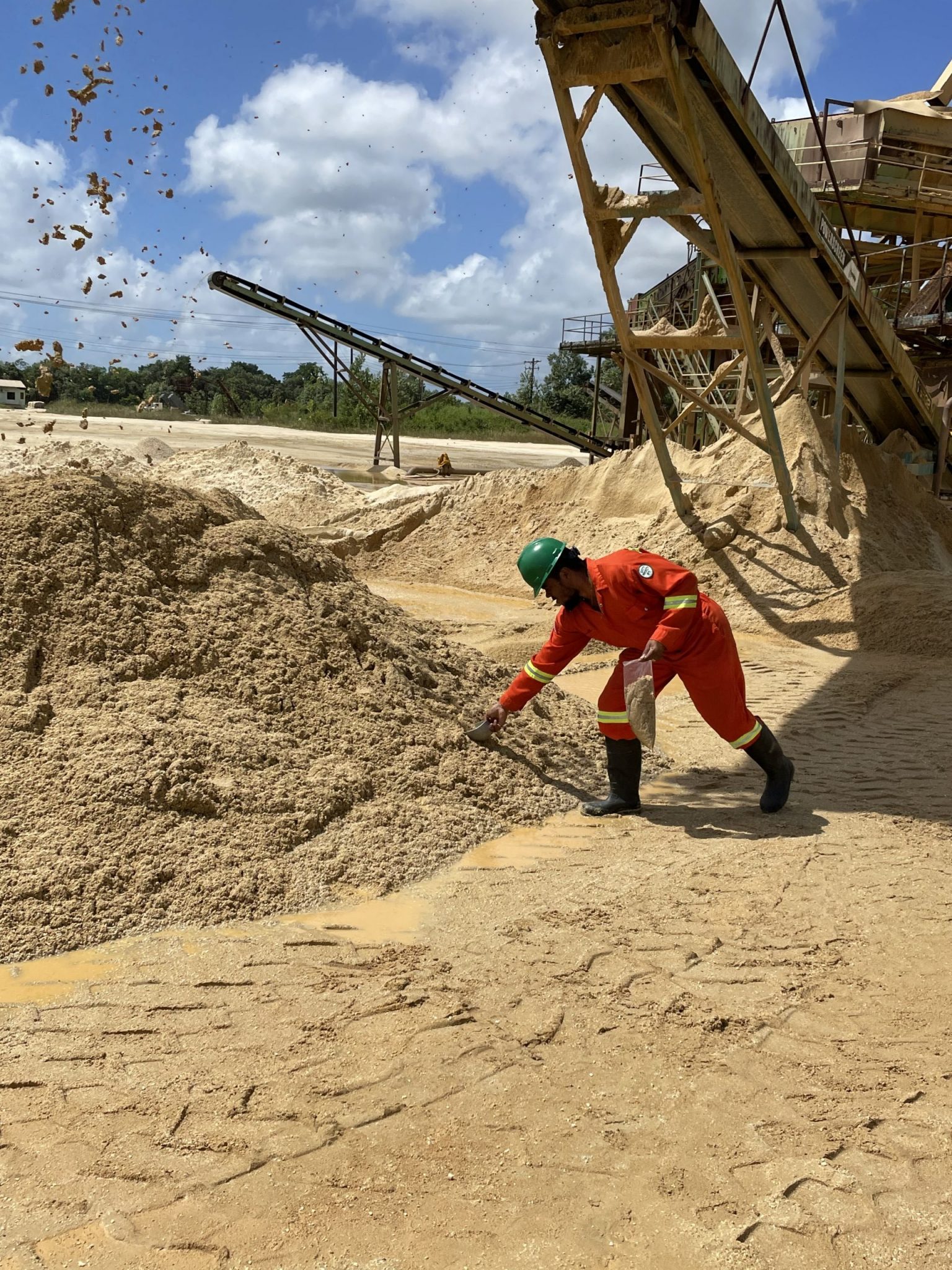 The image showcases an individual in PPE—red overalls, black boots, and a green hard hat—bending down to collect a sample from a sandy surface. Overhead, a conveyor belt system drops freshly processed material onto a large pile, demonstrating active industrial operation. This scene emphasizes the importance of safety, environmental vigilance, and resource management in extractive or processing industries.