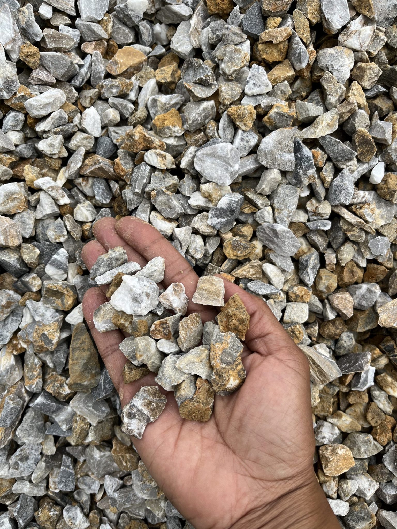This image shows a close-up of medium-sized crushed stones held in an outstretched hand, offering a clear view of the material’s size and texture. The stones are predominantly angular, with hues ranging from white and light gray to rusty brown, suggesting they may be a mix of limestone and iron-stained material. The hand provides a helpful scale reference, indicating the aggregate size is approximately 10–20 mm—often used for drainage, pathways, or base material in construction.