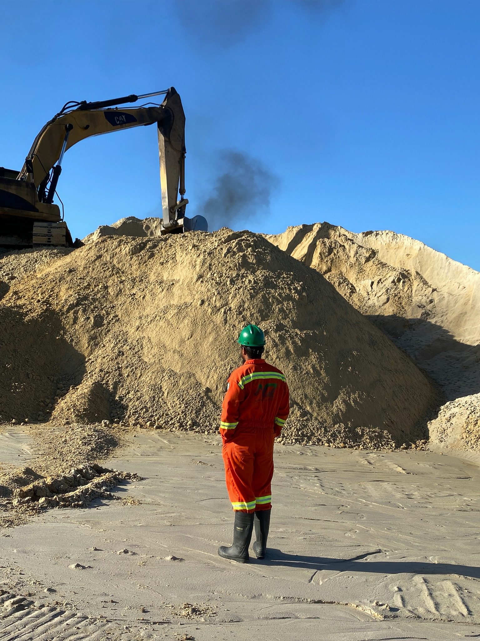 This image captures a field engineer or construction worker in full safety gear—orange coveralls, rubber boots, and a green helmet—standing on a flat sandy area while overseeing a CAT excavator in operation. The heavy equipment is digging into a massive stockpile of silt sand as black exhaust smoke rises into the blue sky, indicating ongoing excavation and material handling activities. The image portrays a blend of active machinery and responsible site supervision.