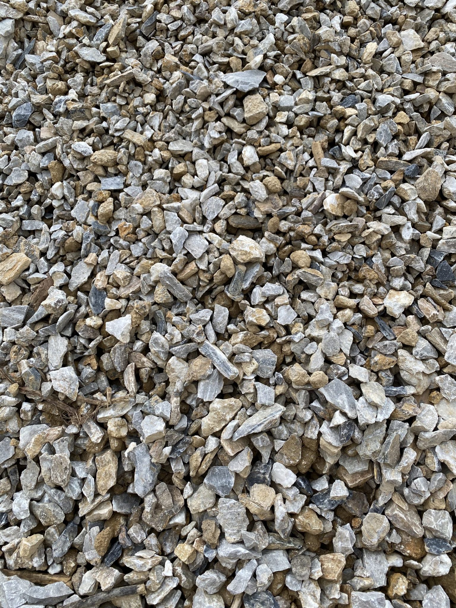 A detailed image showing a mix of crushed stones in a range of sizes and colors, including light gray, beige, tan, and off-white. The stones appear sharp and angular, typical of quarry-processed gravel used in construction, particularly for road base, drainage layers, or concrete work. Fine particles and small debris are visible between the rocks.