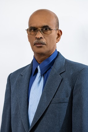 The image features a professional man with a bald head and a neatly trimmed mustache, wearing bronze-framed glasses. He is dressed in a blue suit with a matching dress shirt and a light blue tie. He has a serious and composed expression, standing against a plain light-colored background.