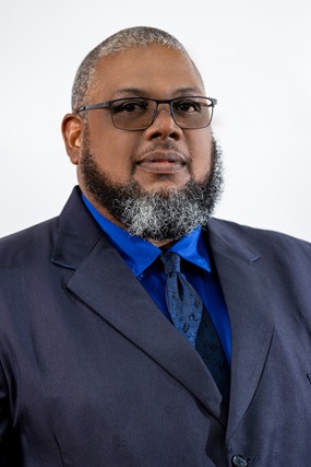 The image features a professional man with short-cropped gray hair, a well-groomed salt-and-pepper beard, and black-framed glasses. He is wearing a dark navy suit, a vibrant blue dress shirt, and a patterned navy tie. He has a confident and composed expression, standing against a plain light-colored background.