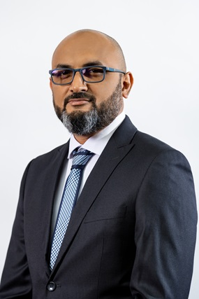 The image shows a professional man with a shaved head, a well-groomed salt-and-pepper beard, and black-framed glasses. He is wearing a dark suit, a white dress shirt, and a blue patterned tie. He has a confident and composed expression, standing against a plain light-colored background.