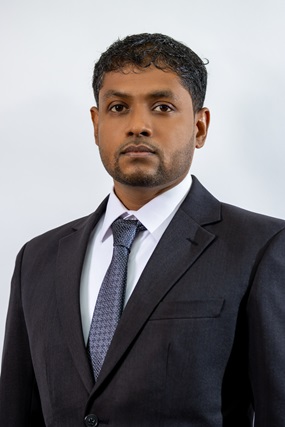 The image features a professional man with short, curly dark hair and a neatly groomed beard. He is wearing a dark suit, a white dress shirt, and a patterned grey tie. He has a serious and confident expression, standing against a plain light-colored background.