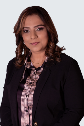 The image features a professional woman with shoulder-length, wavy brown hair. She is dressed in a black blazer over a satin-striped blouse in shades of pink and black. She has a poised and confident expression, standing against a light-colored background.