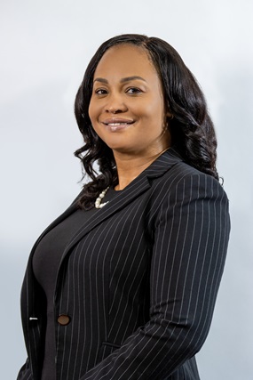 The image shows a professional woman with wavy black hair wearing a black pinstripe blazer and a black top. She is smiling confidently, standing against a plain light-colored background.