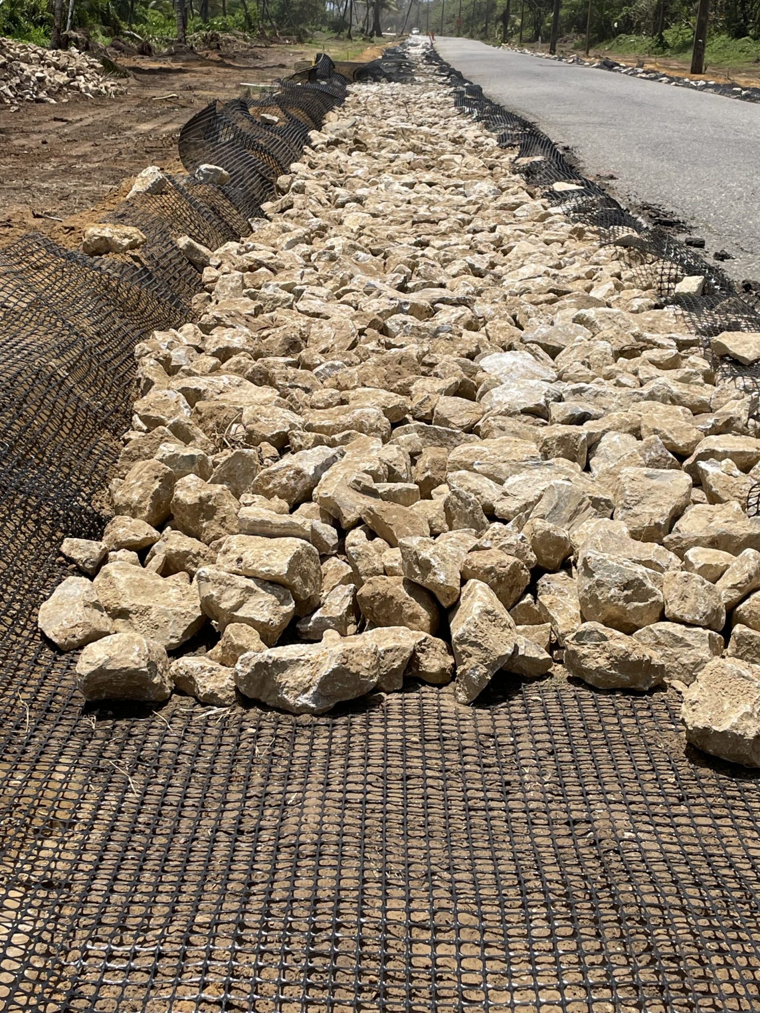 The image shows a row of large rocks laid on black netting next to a road. This setup, called riprap, helps stop the soil from washing away and keeps the roadside strong.