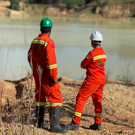 Two workers in orange safety gear and helmets stand near a quarry pond, observing the water and surrounding landscape.