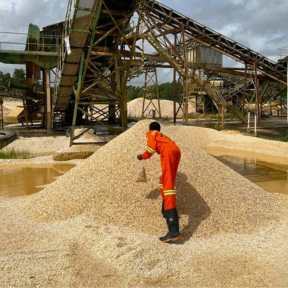 Dressed in full safety gear, a technician at National Quarries is seen collecting samples from a freshly processed pile of aggregate beneath an active conveyor system.