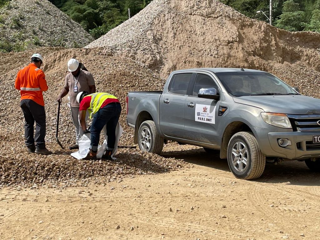 Set in a rugged quarry environment, the image depicts three individuals in safety vests and helmets engaged in the inspection and sampling of gravel. They are positioned next to a grey Ford pickup truck labeled with the PURE Unit insignia, part of the Ministry of Works and Transport. Behind them are large mounds of aggregate, highlighting the scale of material handling at the site.