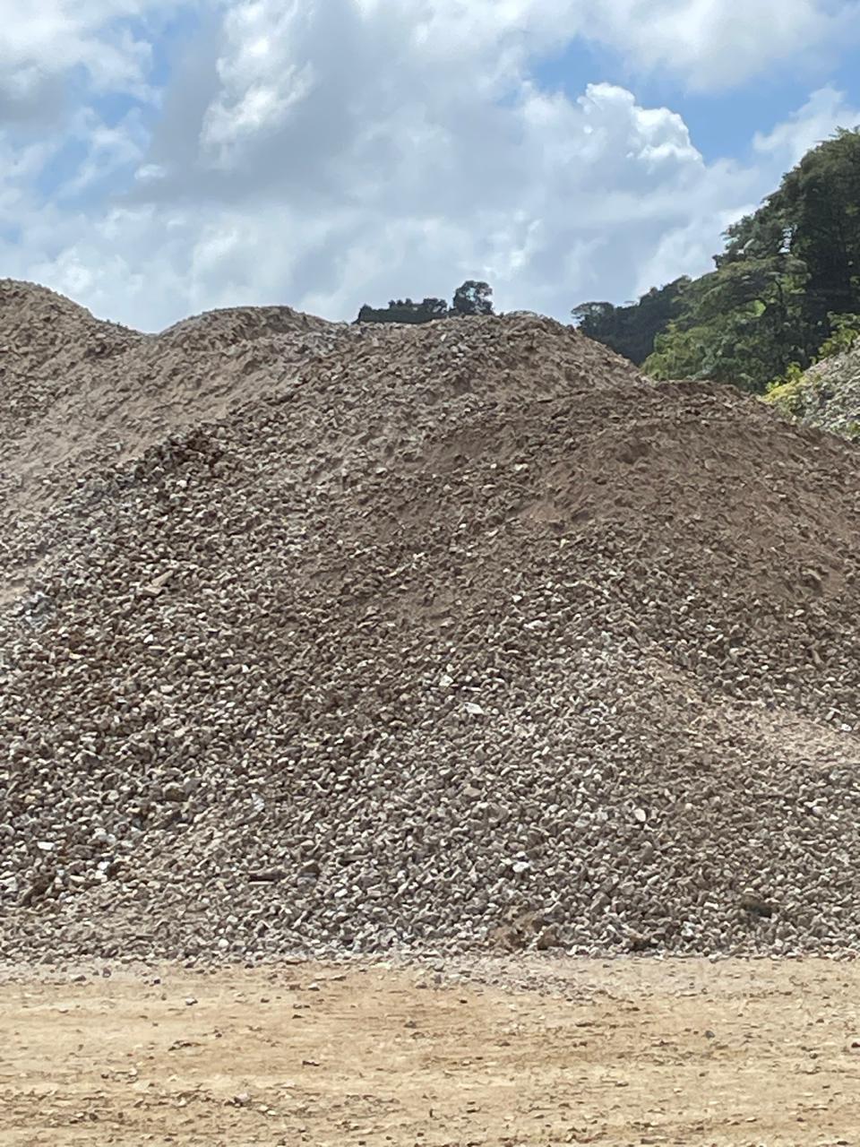 The image shows a large mound of crushed stone and gravel at an outdoor quarry or construction material site. The material is used in road construction, foundation work, and other infrastructure projects. A backdrop of trees and a partly cloudy sky can be seen, highlighting the open nature of the site.