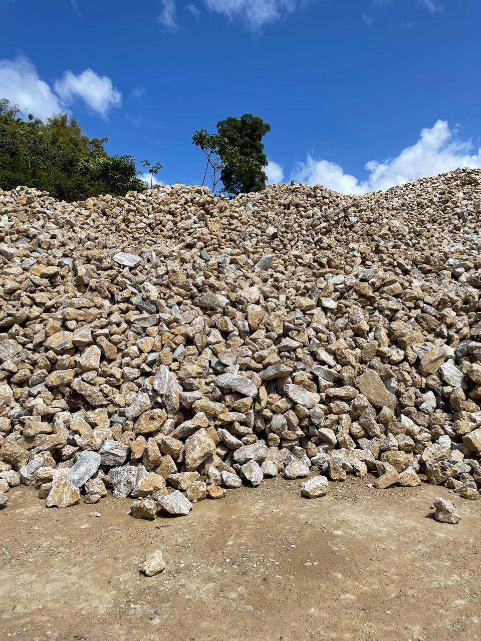 A large mound of crushed rocks under a bright blue sky, with green trees in the background.