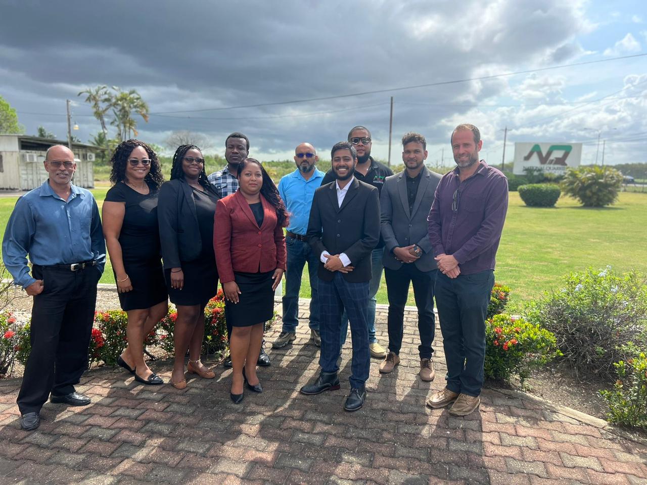 A group of ten professionally dressed individuals pose outdoors in front of a manicured lawn and the “National Quarries” sign, with dark storm clouds looming in the background.