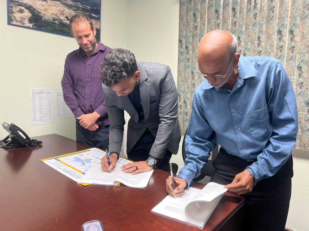 In an indoor office setting, two men are seen signing official documents on a desk while another stands behind them, observing. The atmosphere is professional, with the table holding a folder, printed documents, and a telephone. A map and documents on the wall suggest the nature of the agreement may be related to geography or land use. The collaboration appears formal and significant, marked by mutual understanding and cooperation