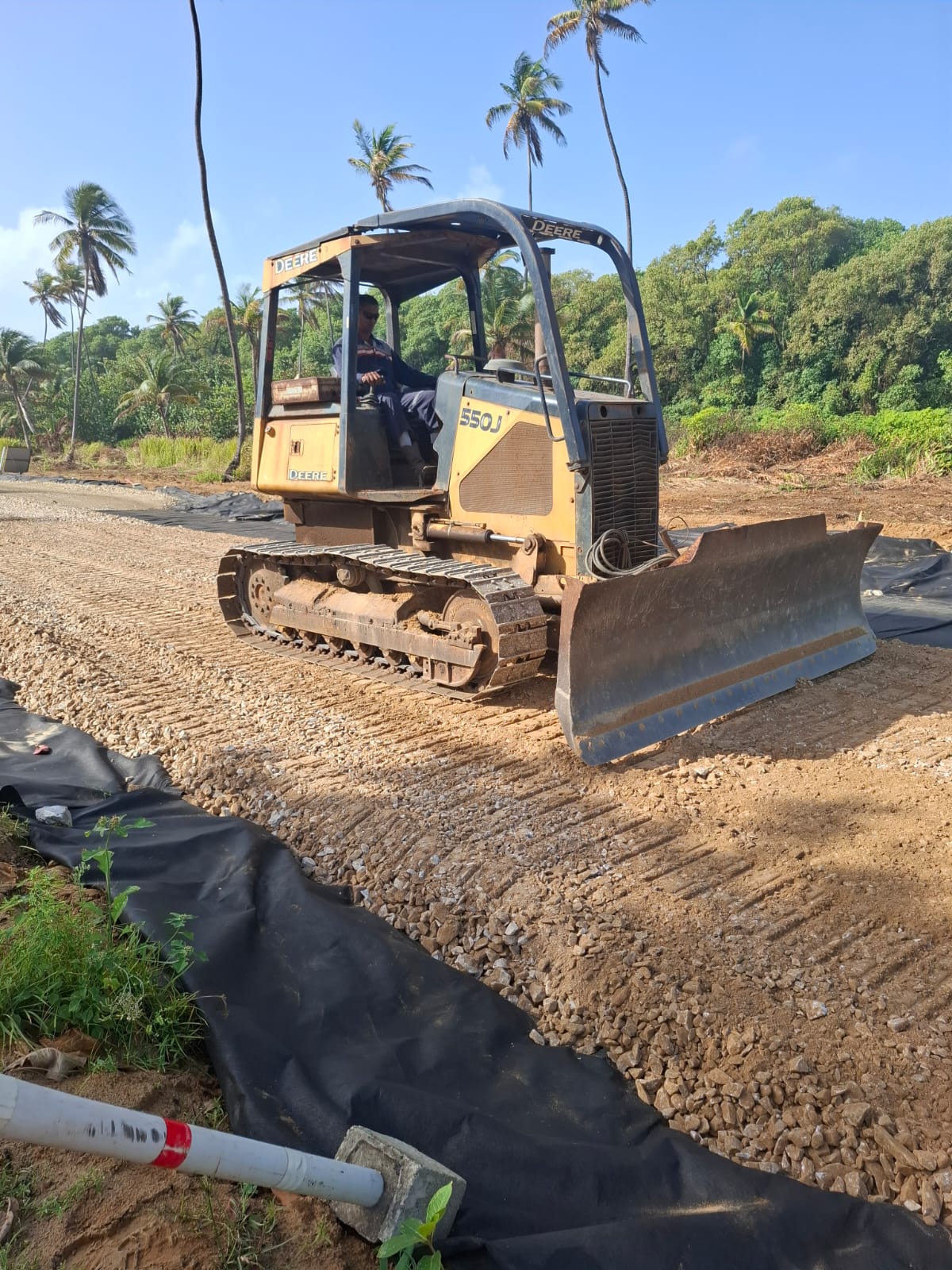The image shows a John Deere 550J bulldozer operated by a worker, spreading and leveling gravel on top of black geotextile fabric. This process is part of roadbed or foundation preparation in a tropical setting with palm trees and dense greenery in the background. The geotextile helps improve ground stability and drainage.